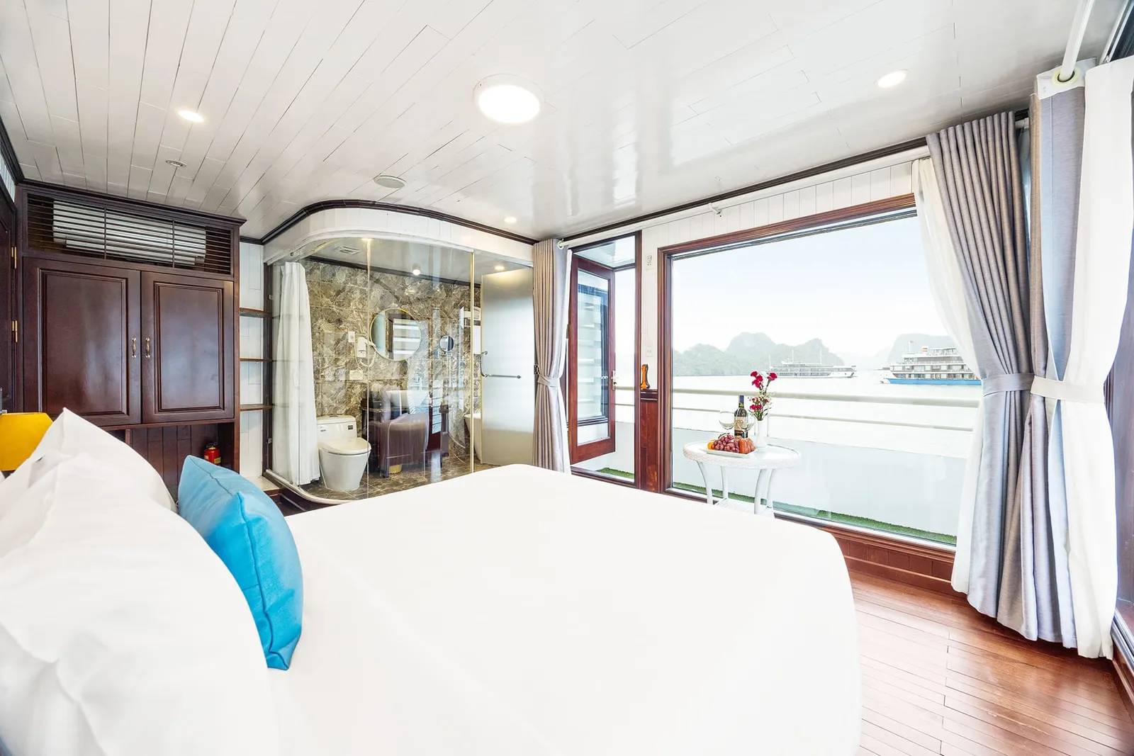 A cruise cabin with a white bed and blue cushion, a glass-walled bathroom, and a large window and balcony overlooking calm water, limestone karsts, and another cruise boat.
