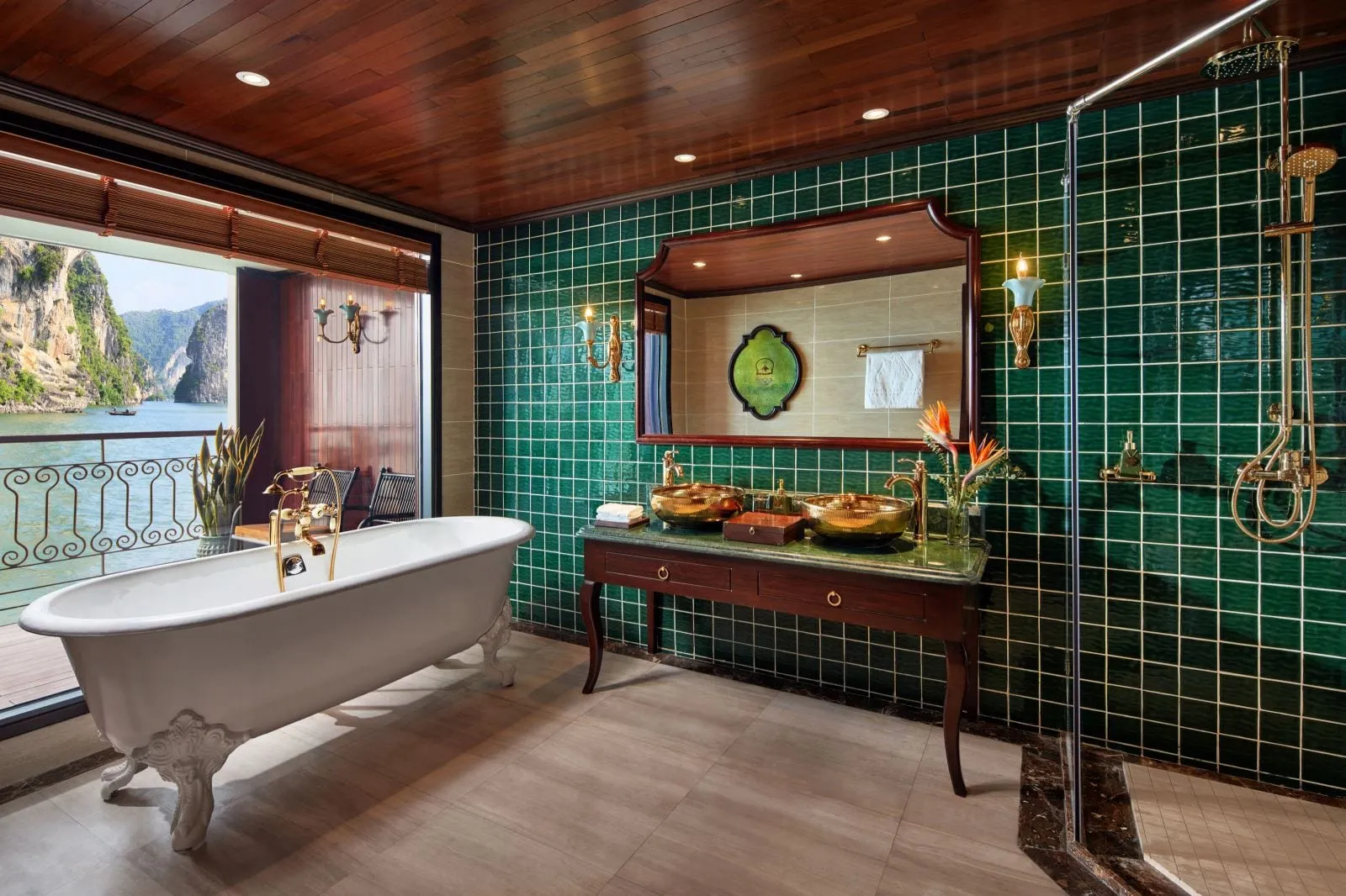 A bathroom with green tiled walls, a white clawfoot tub, brass fixtures and gold basins on a wooden vanity, and an open balcony door showing limestone karsts rising from calm green water.