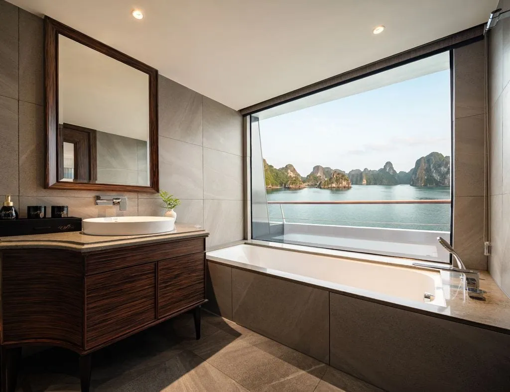 A cruise ship bathroom with a wooden vanity, mirror, and built-in bathtub beside a large window overlooking calm water and limestone karst islands, with small branded amenity items on the counter.