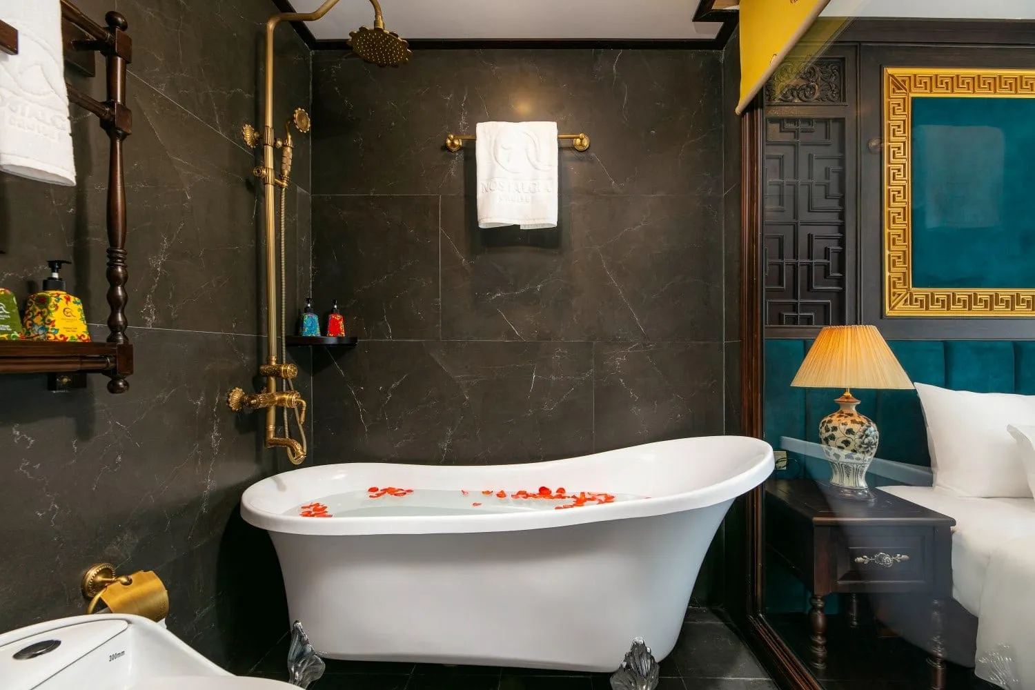 A dark marble bathroom with a white freestanding clawfoot tub holding red petals, brass shower fixtures, and a towel embroidered with 'Nostalgia', next to a glass-partitioned bedroom with teal headboard and a lamp.