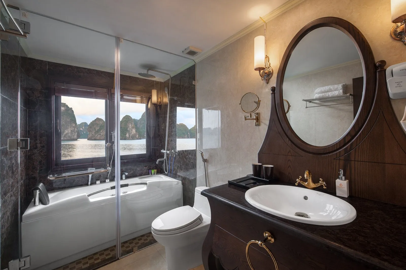 An en-suite bathroom with a dark wood vanity, oval mirror, white bathtub and toilet, with a window showing limestone karst islands over calm water.