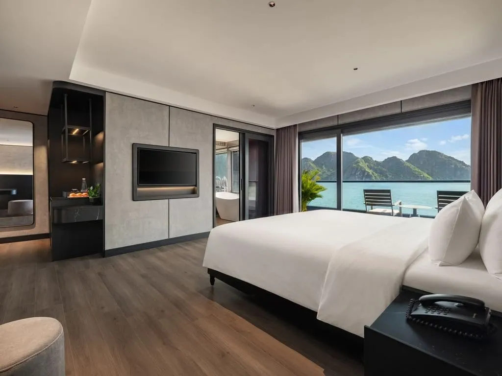 A modern cruise cabin with a large white bed, wall-mounted TV, bathtub and a balcony with chairs overlooking turquoise water and green limestone karst hills.