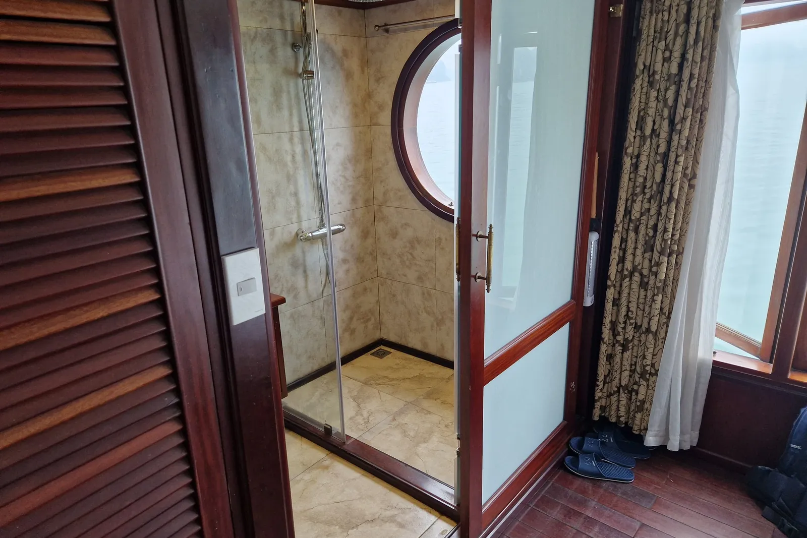 A boat cabin interior with a glass-enclosed shower, round porthole window, wooden furnishings, curtains, and a view of water through windows.