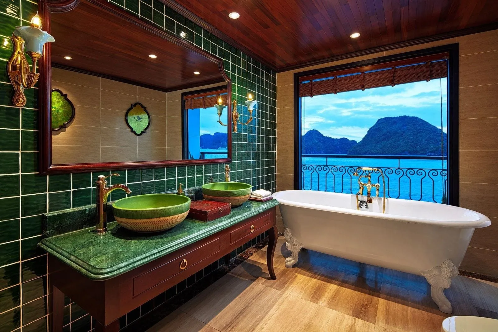 A bathroom with green tiles, green marble vanity with two vessel sinks, a white clawfoot tub, and a large window showing blue water and karst islands.