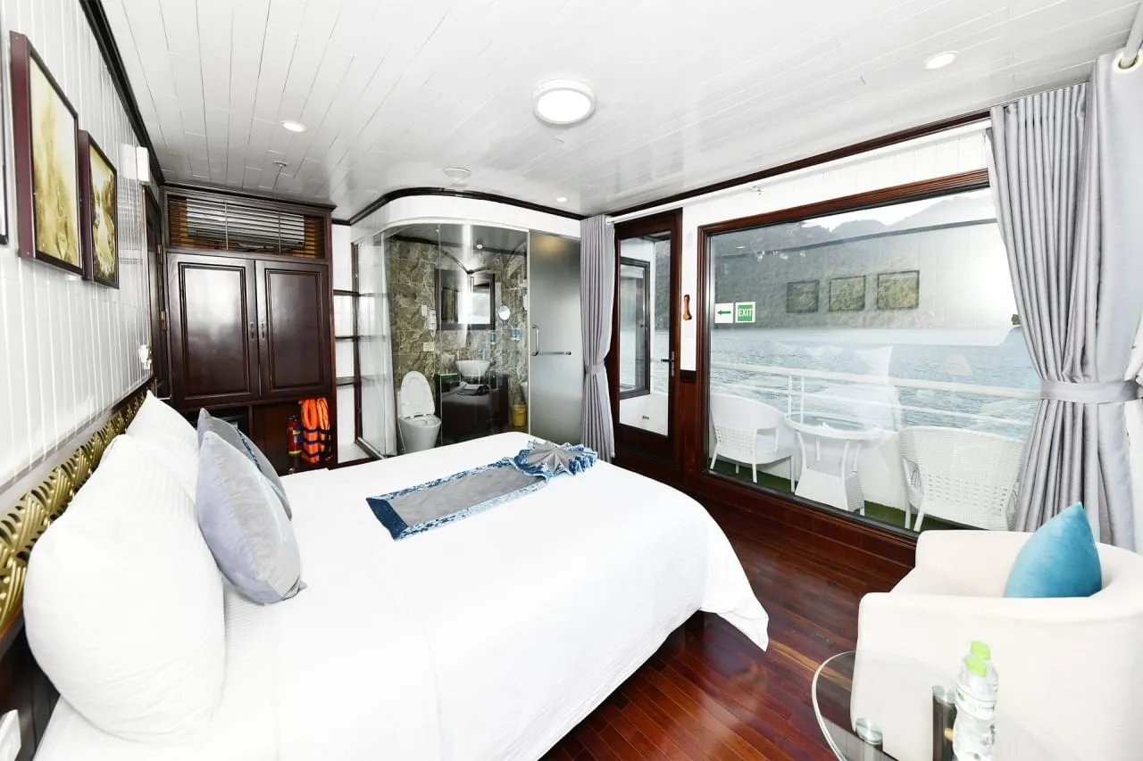 A cruise cabin with a white double bed, glass-walled bathroom, wooden wardrobe with life jackets, and a large window showing a private balcony with white chairs and a table overlooking water and limestone karst hills.