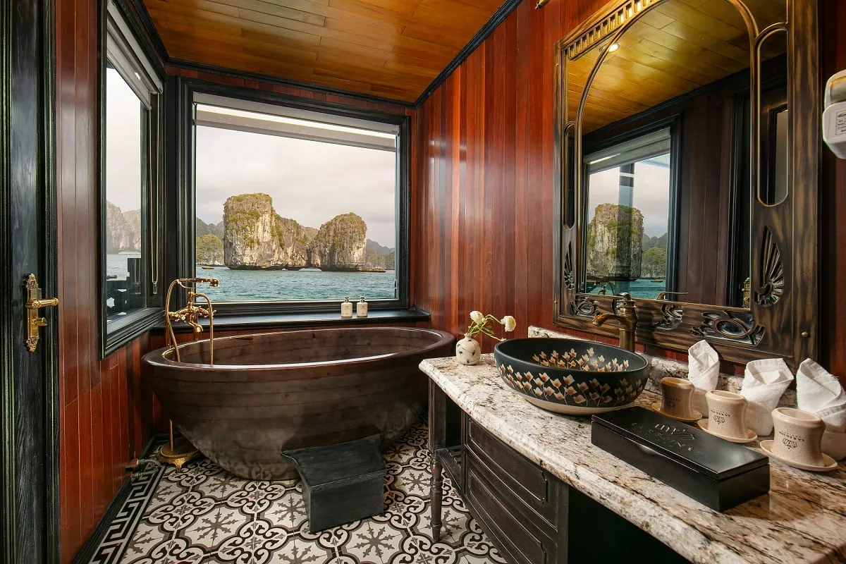A luxury cruise cabin bathroom with wooden bathtub, decorative sink, and large windows showing limestone karsts rising from blue-green water.