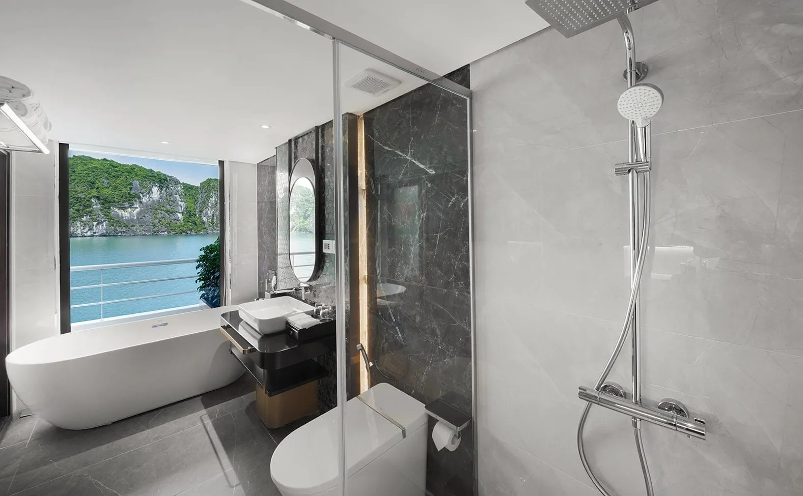 A modern marble bathroom with a freestanding tub, vanity, toilet and rain shower, with a window showing a green limestone karst and calm bay water beyond a railing.