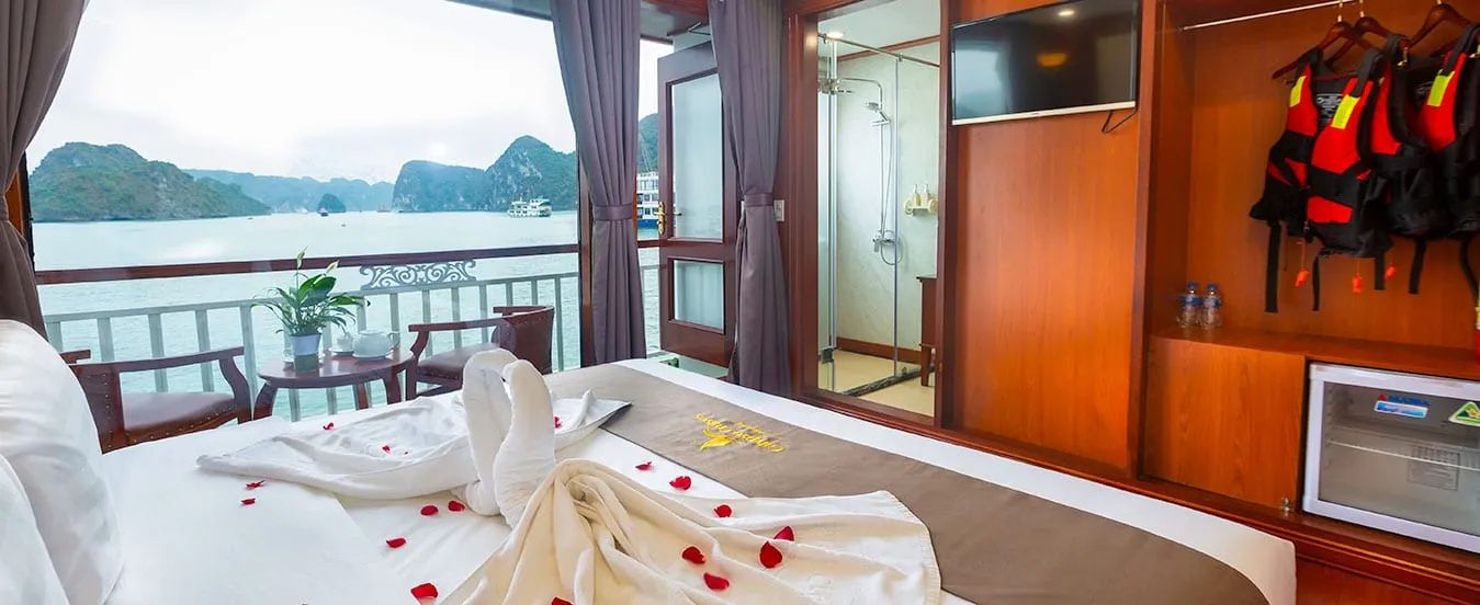 A cruise cabin interior with a bed decorated with towel swans and rose petals, open balcony doors showing karst limestone islands and water, life jackets hanging on the wall, and wood-paneled walls with a glass shower visible.