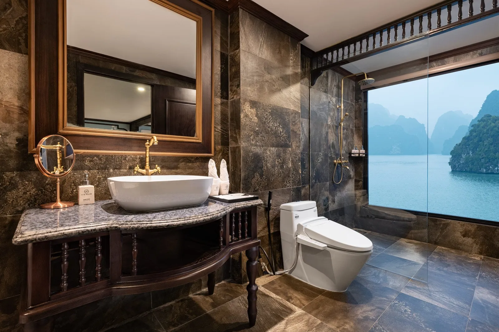 A dark marble ensuite bathroom with a vessel sink on a wooden vanity, a toilet, and a rain shower beside a large window overlooking limestone karst islands and calm water.