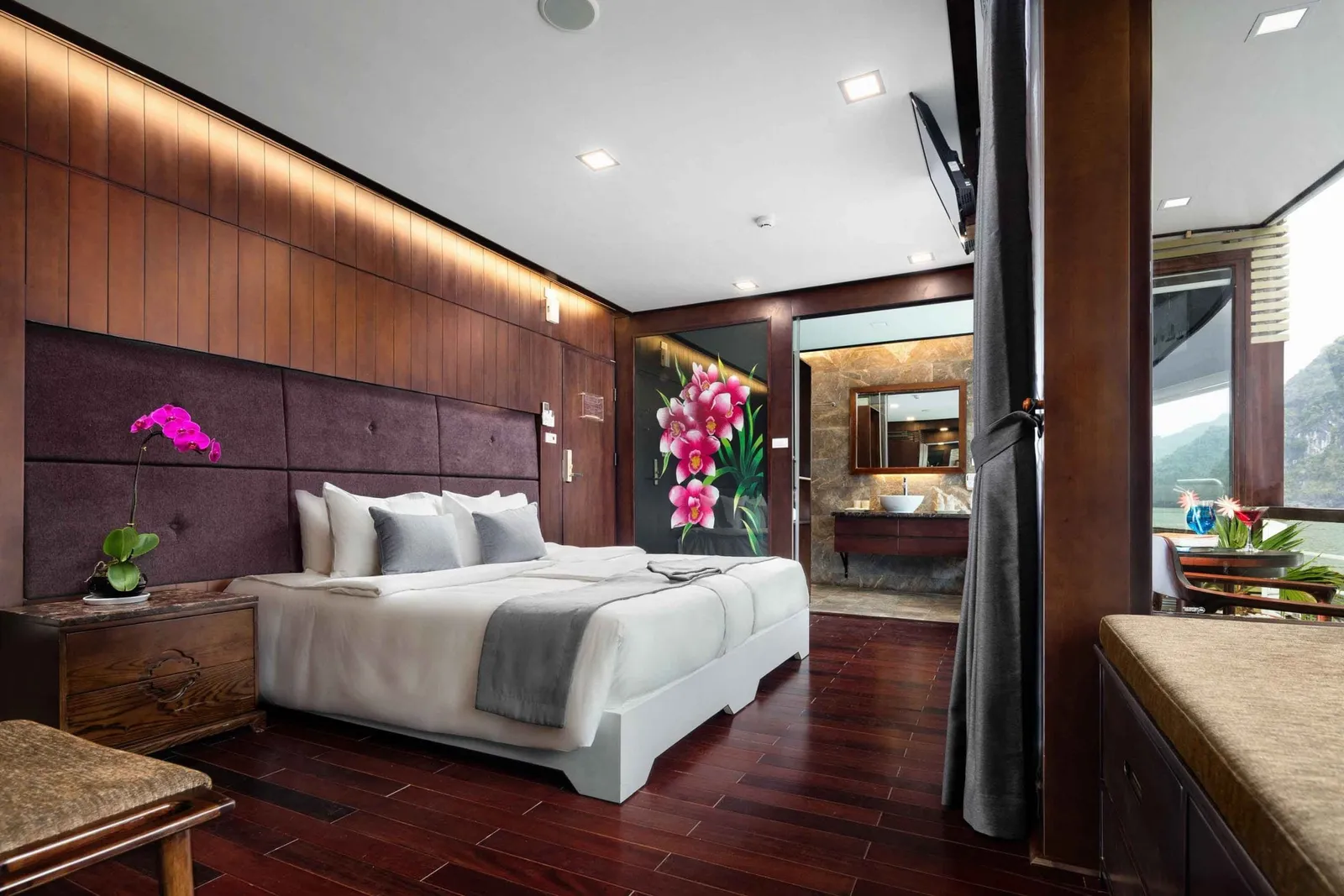 A dark-wood cruise cabin with a white double bed, orchid decor, an ensuite bathroom behind, and a balcony with limestone karst hills visible through the window.