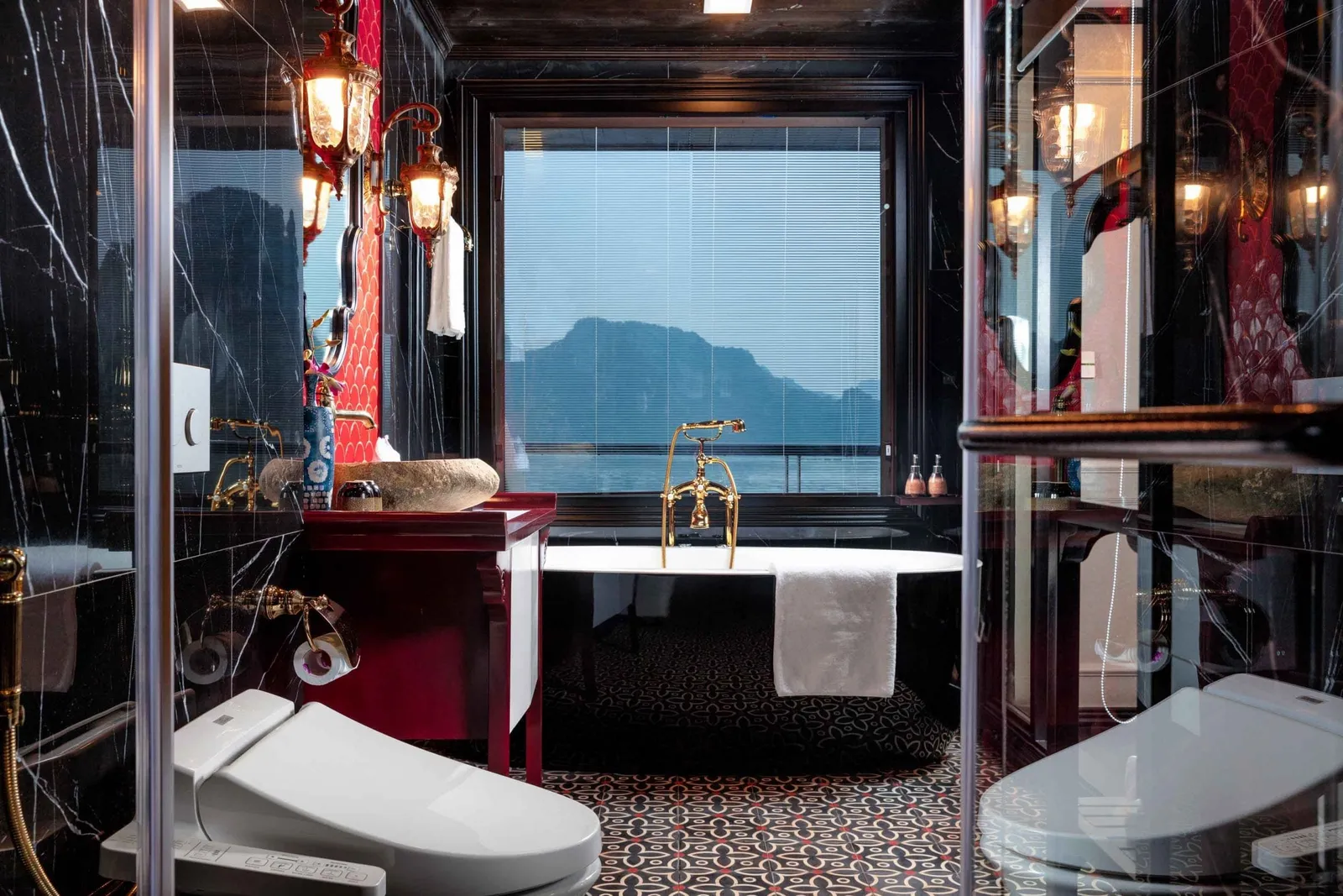A luxury bathroom with black marble walls, red vanity, gold fixtures, freestanding tub and bidet toilet, with a window showing misty limestone karst hills over the water.