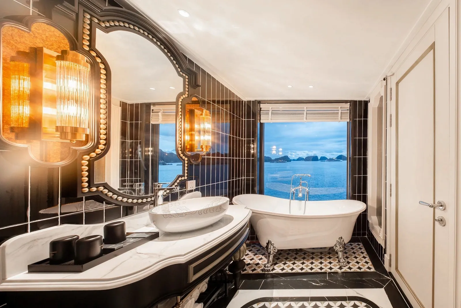 A luxurious black-and-white marble bathroom with a freestanding clawfoot tub and ornate mirror, with a large window looking out onto a bay dotted with limestone karst islands at dusk.