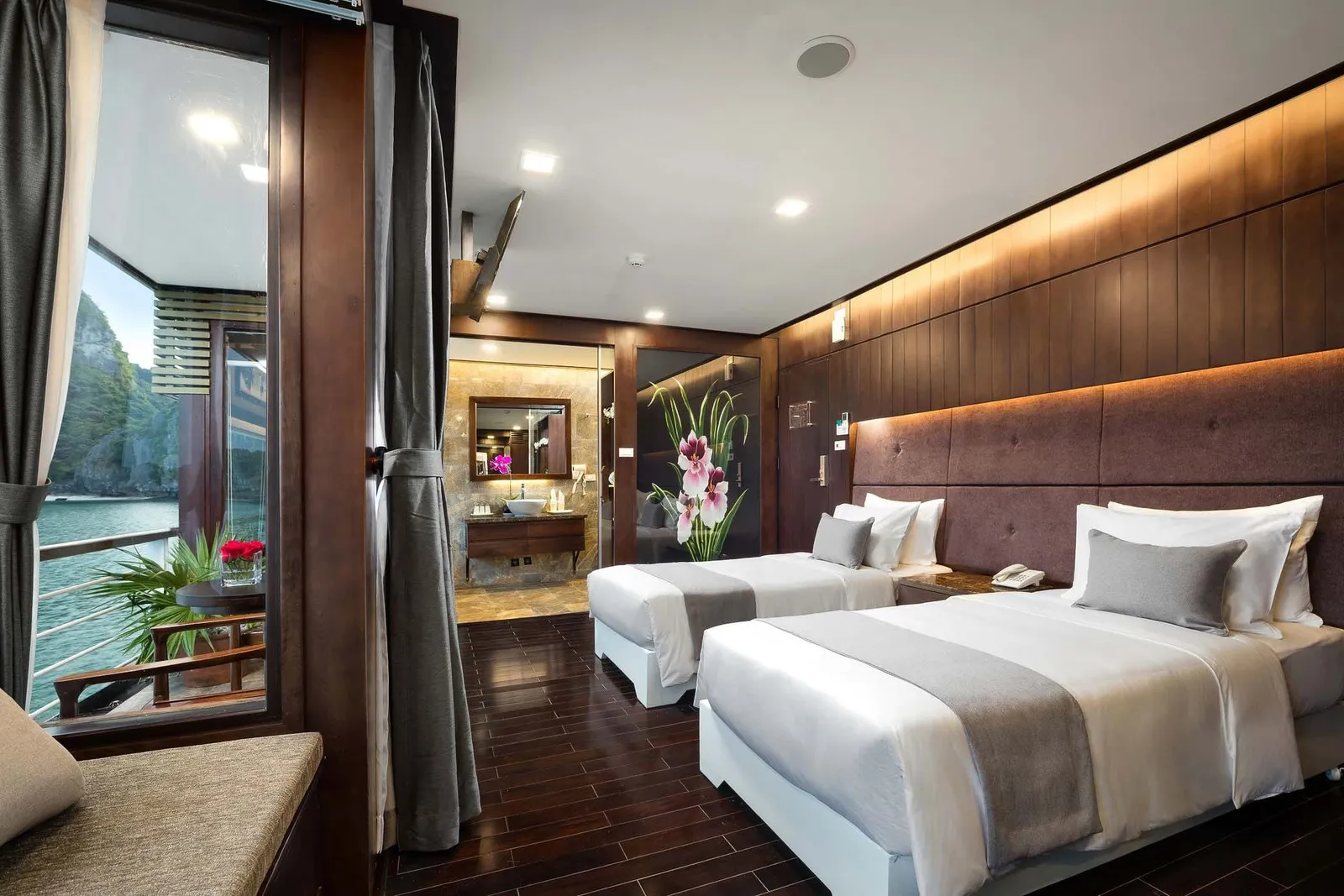 A cabin with two twin beds, dark wood floor and paneling, and an ensuite bathroom, with a balcony door showing calm water and limestone karst cliffs outside.