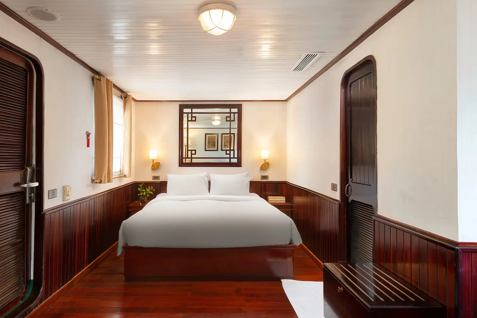 A cabin with a double bed in white linens, dark red-brown wood paneling and floor, a latticed mirror, wall lamps, a window with tan curtains, and louvered wooden doors on both sides.