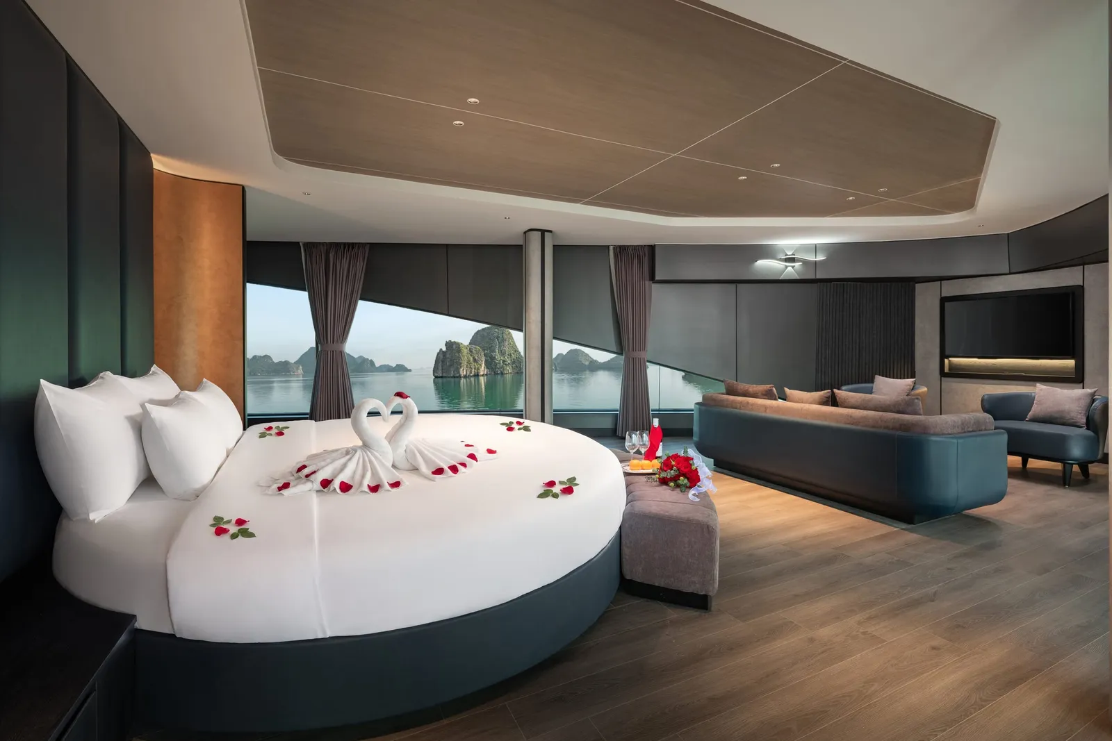 A luxury cabin with a round white bed decorated with towel swans and rose petals, a sofa, armchair and TV, and wide windows looking out onto calm water and limestone karst islands.