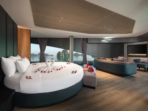 A luxury cabin with a round white bed decorated with towel swans and rose petals, a sofa, armchair and TV, and wide windows looking out onto calm water and limestone karst islands.