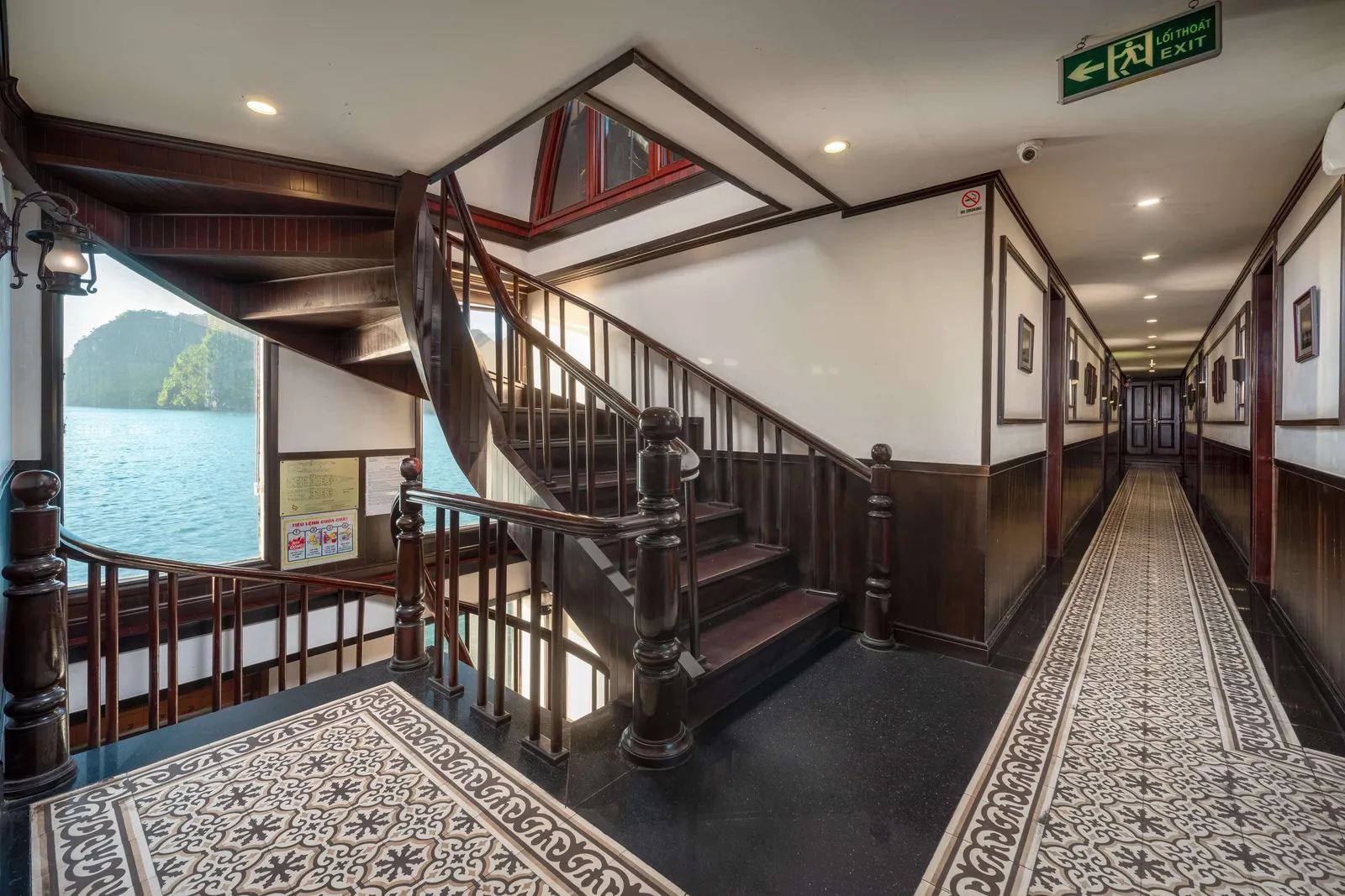 Interior hallway and wooden staircase of a cruise boat with exit signage, patterned carpet, and a view of karst islands through an open doorway.