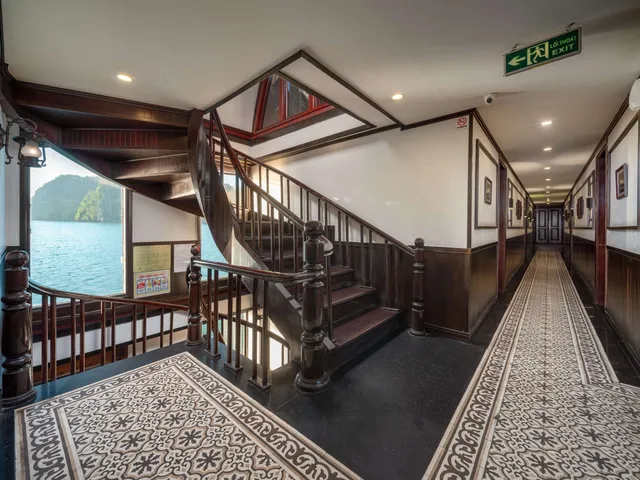 Interior hallway and wooden staircase of a cruise boat with exit signage, patterned carpet, and a view of karst islands through an open doorway.