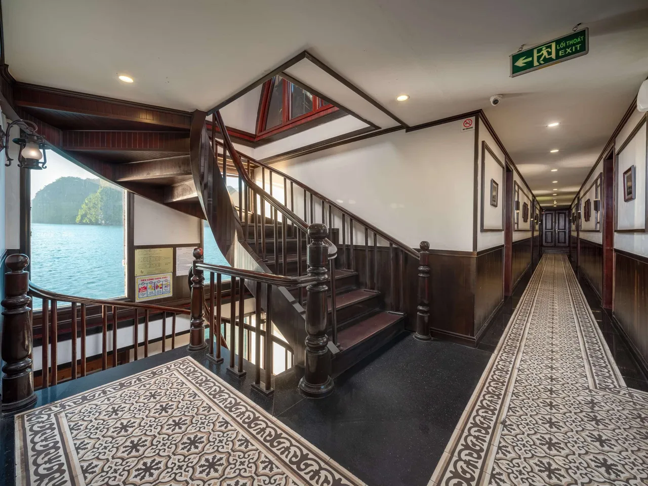 Interior hallway and wooden staircase of a cruise boat with exit signage, patterned carpet, and a view of karst islands through an open doorway.