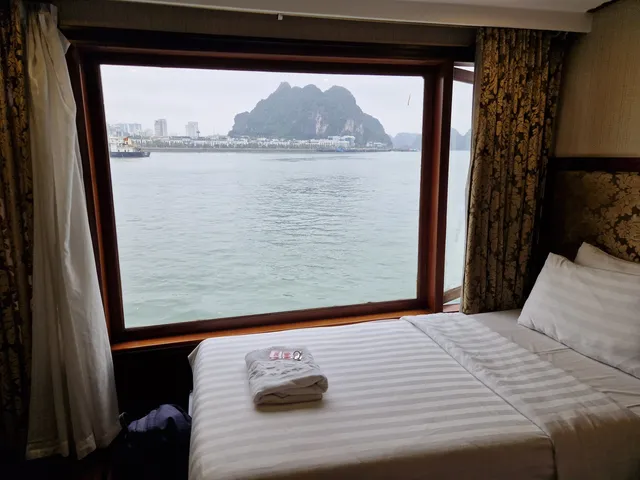 A boat cabin interior with a bed and large window showing a limestone karst island and waterfront buildings across the water.