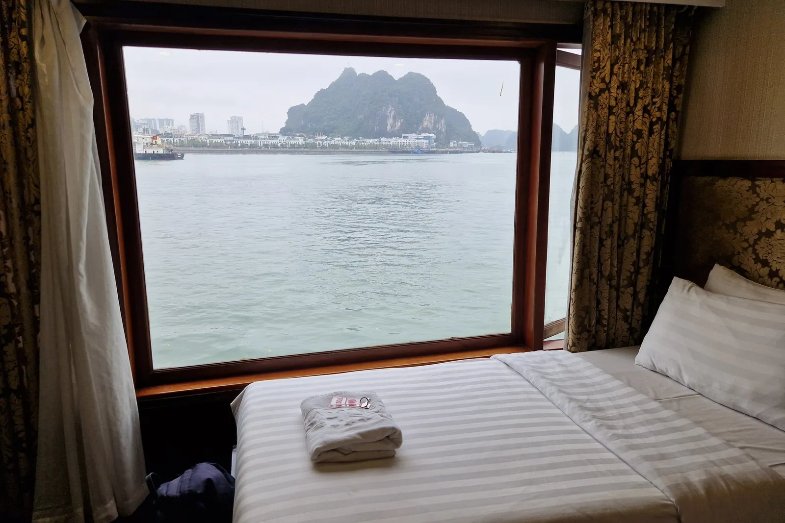 A boat cabin interior with a bed and large window showing a limestone karst island and waterfront buildings across the water.