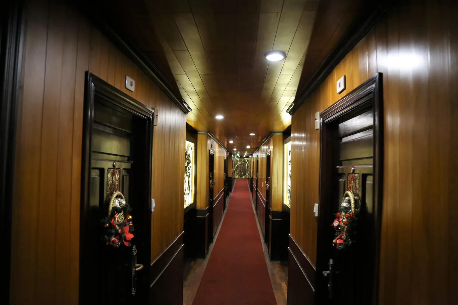 A long interior corridor with wood paneling, black cabin doors decorated with floral ornaments, red carpet runner, and recessed ceiling lights.