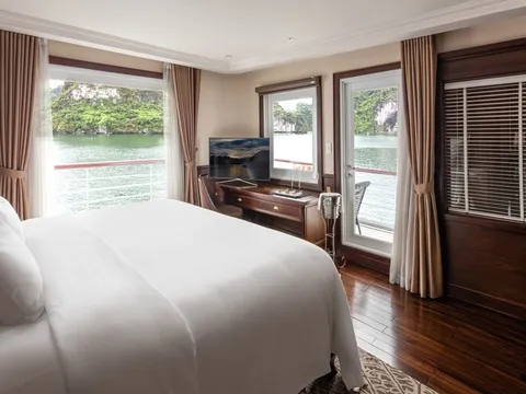A cruise ship cabin with a white-draped bed, wood desk with TV, and brown curtains, with windows and a balcony door looking out over green water and limestone karst islands.