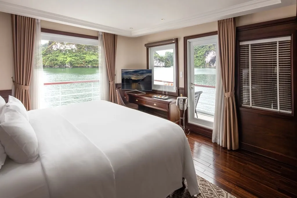 A cruise ship cabin with a white-draped bed, wood desk with TV, and brown curtains, with windows and a balcony door looking out over green water and limestone karst islands.
