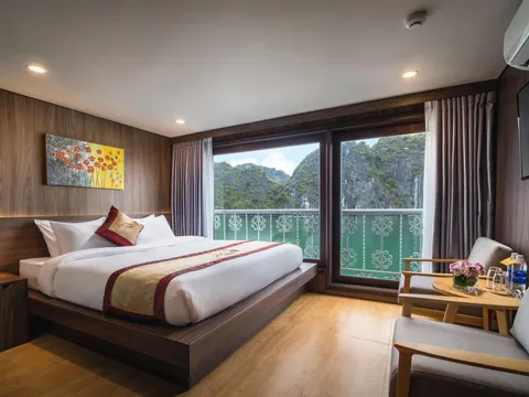 A wood-paneled cabin with a double bed, a branded cushion on it, two armchairs and a small table, and balcony doors opening onto green water and limestone karst peaks.
