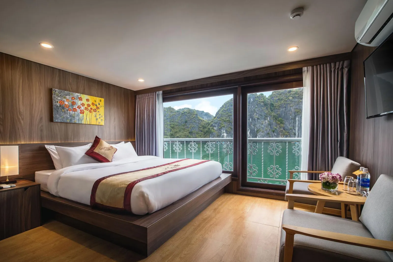 A wood-paneled cabin with a double bed, a branded cushion on it, two armchairs and a small table, and balcony doors opening onto green water and limestone karst peaks.