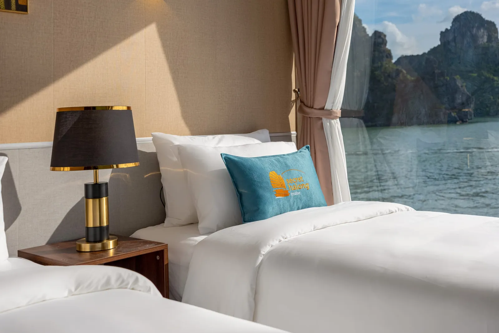 A cabin with two twin beds in white linen, teal cushions, bedside lamps and a window with limestone islands outside.