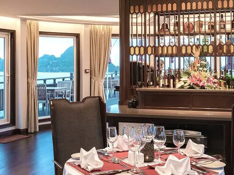 A boat dining room with set tables, wooden bar with flowers and bottles, and large windows showing calm water, karst hills and a sun deck with loungers.