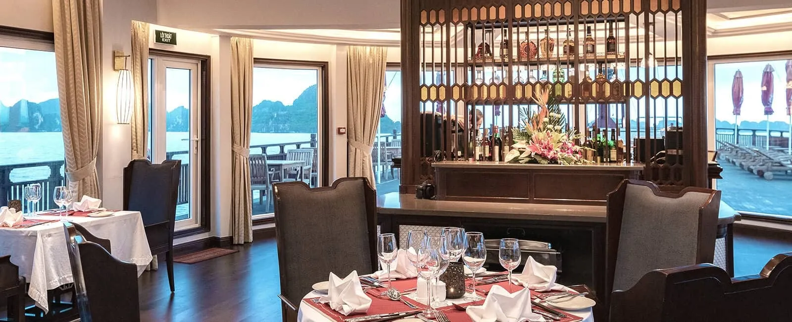 A boat dining room with set tables, wooden bar with flowers and bottles, and large windows showing calm water, karst hills and a sun deck with loungers.