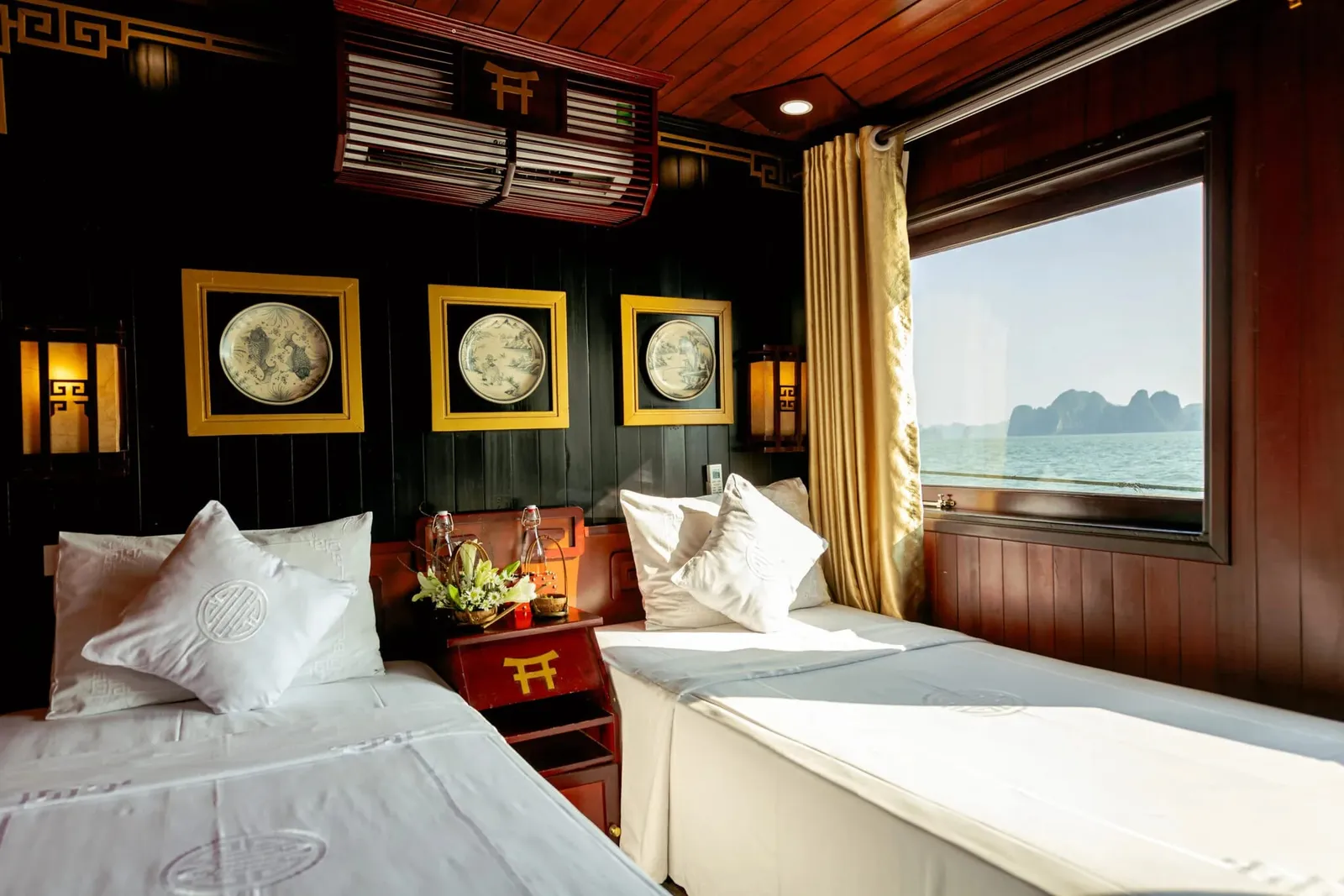 A twin-bed wooden cabin with dark paneled walls, framed decorative plates, and a window overlooking calm water and limestone karst islands, with embroidered emblems on the pillows and linens and gold torii-style symbols on the bedside table and wall unit.
