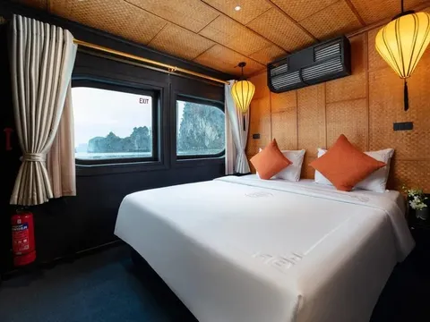 A cabin with a white double bed, orange cushions, woven bamboo walls and silk lanterns, with porthole windows showing limestone karsts over misty water, a fire extinguisher and an EXIT sign, and a subtle embroidered pattern on the bed linen.