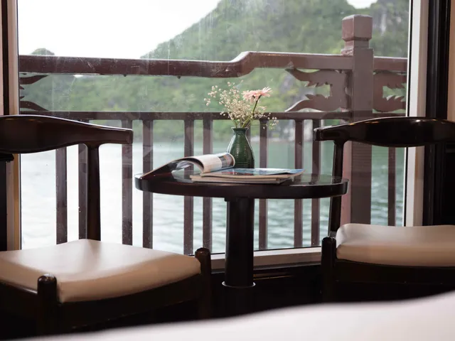 Two dark wooden chairs with cream cushions flank a small round table holding an open magazine and a green vase of flowers, in front of a window overlooking a wooden balcony railing, calm water and a green karst hill.