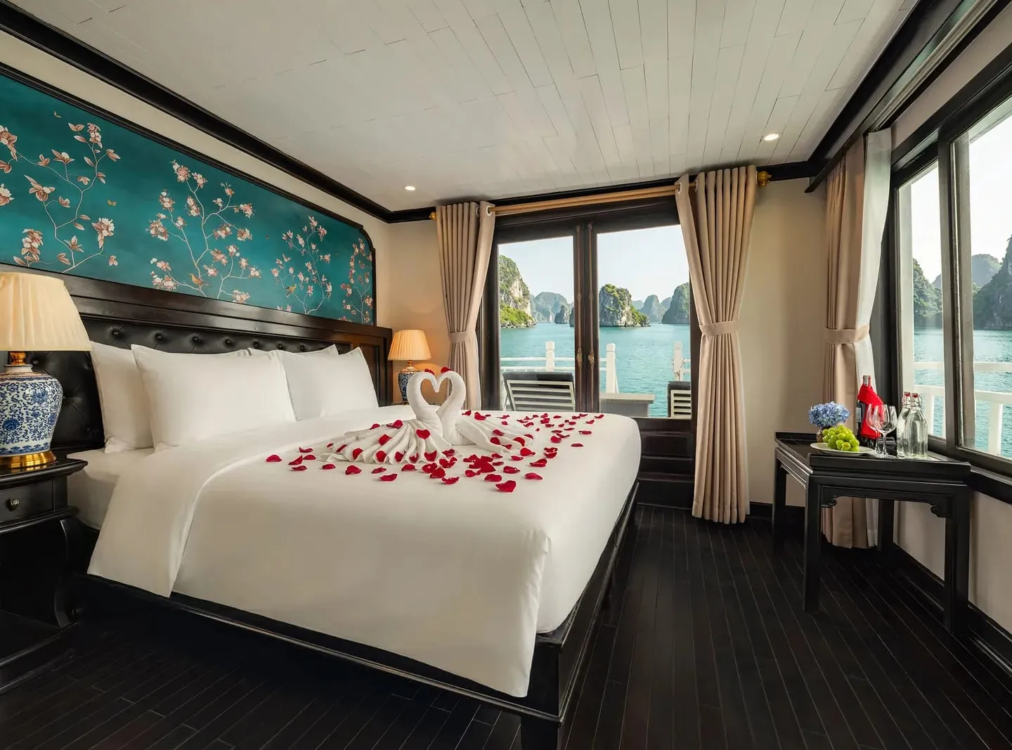 A cruise cabin with a white double bed decorated with a towel-swan heart and red rose petals, a teal floral headboard panel, and glass doors opening onto a balcony with limestone karst islands in the bay beyond.
