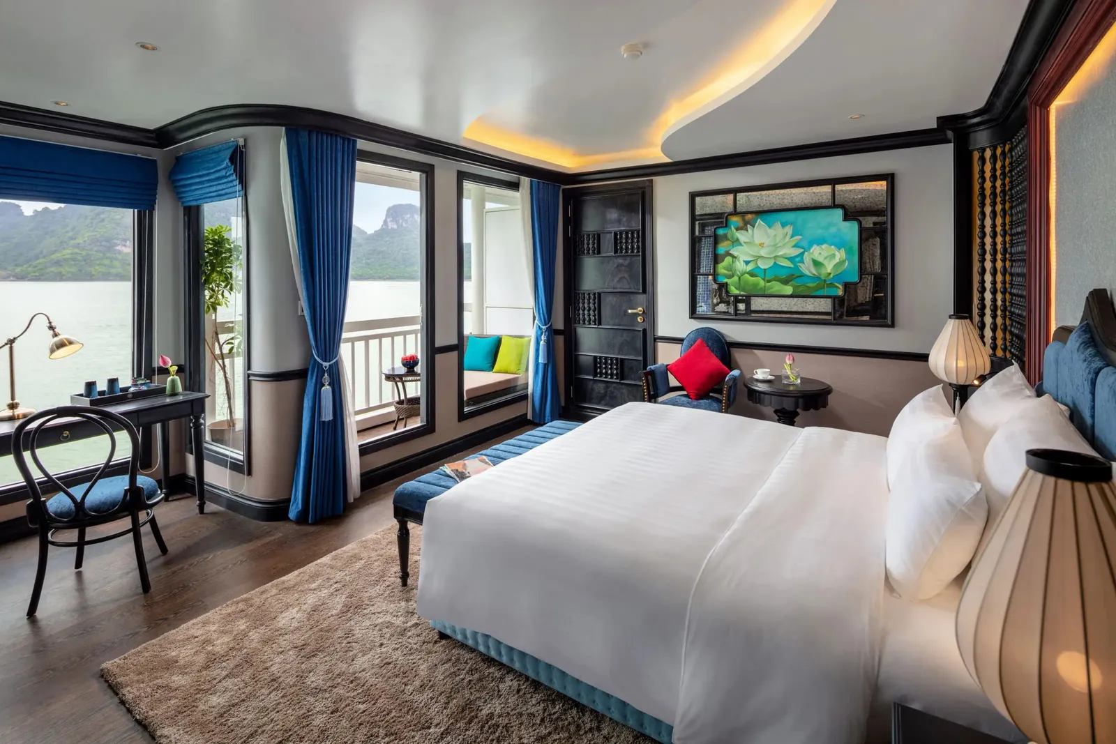 A cabin with a large white bed, blue curtains, a desk and a private balcony, with floor-to-ceiling windows looking onto calm water and limestone karst islands.