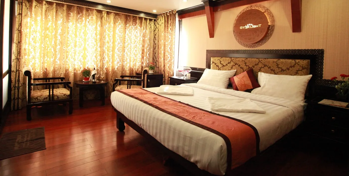 A cabin with a double bed with striped white linen and a rust-colored runner, gold damask curtains, wooden armchairs, a polished wood floor, and a round carved 'Starlight' plaque on the wall above the headboard.
