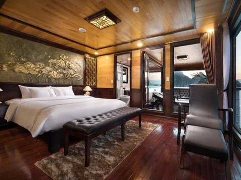 A wood-paneled cruise cabin with a white-bedded king bed, leather bench, lotus relief panel, armchair and ottoman, with a private balcony and a window overlooking limestone karst islands and calm water at dusk.