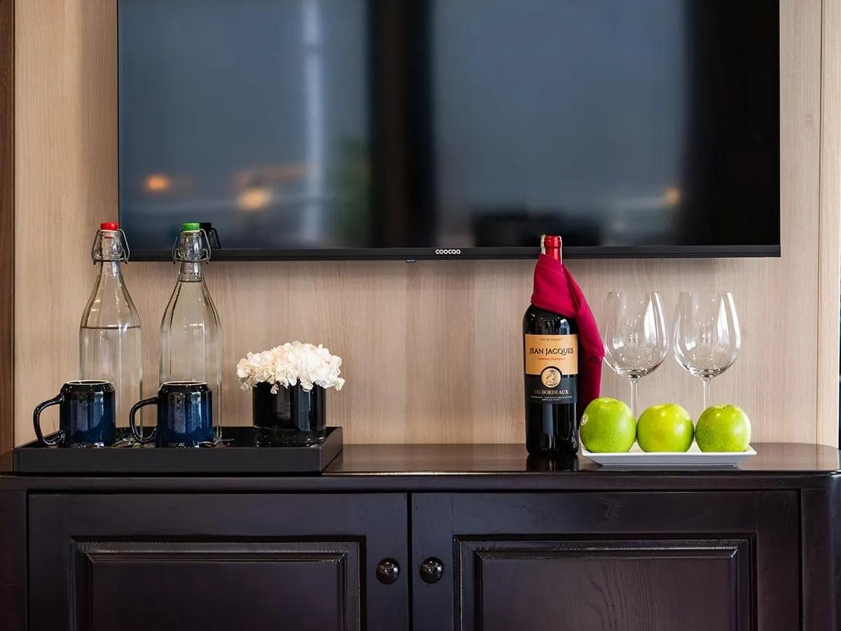 A dark wooden sideboard under a wall-mounted TV holds two glass water bottles, blue mugs on a tray, white flowers, a wine bottle with a red napkin, two wine glasses and three green apples.