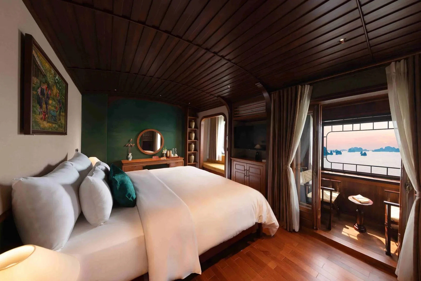 A wood-paneled cabin with a large white-bedded double bed, green accent wall, vanity with round mirror, and a window and balcony with chairs opening onto a bay of limestone karsts at sunset.