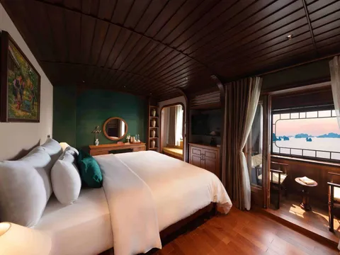 A wood-paneled cabin with a large white-bedded double bed, green accent wall, vanity with round mirror, and a window and balcony with chairs opening onto a bay of limestone karsts at sunset.