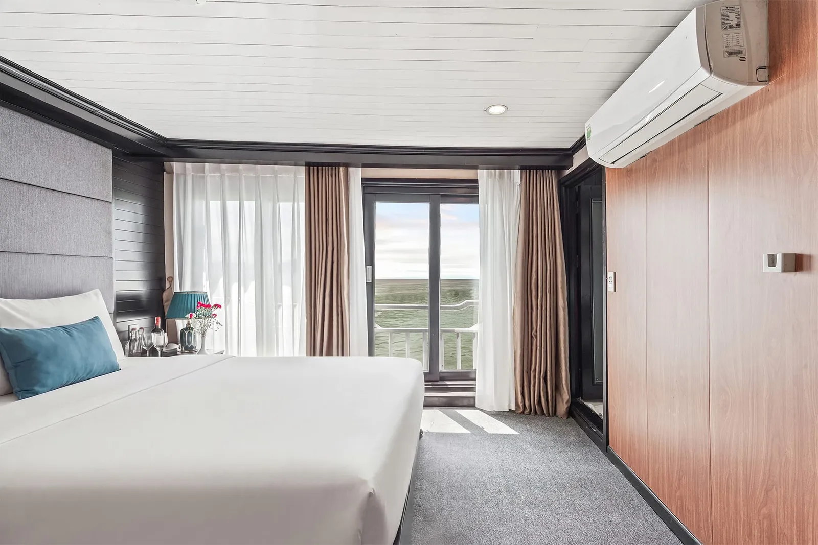 A cabin with a white double bed, teal cushion and lamp, wood-panelled wall with a wall-mounted air conditioner, and sheer and brown curtains beside a glass balcony door that opens onto a railing and a flat expanse of water or green land under a pale sky.