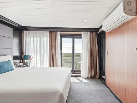 A cabin with a white double bed, teal cushion and lamp, wood-panelled wall with a wall-mounted air conditioner, and sheer and brown curtains beside a glass balcony door that opens onto a railing and a flat expanse of water or green land under a pale sky.