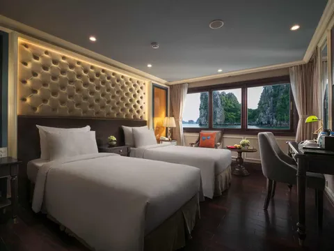 A cruise cabin with two twin beds, a tufted lit headboard, a desk and chair, and windows showing green limestone karsts rising from the water.