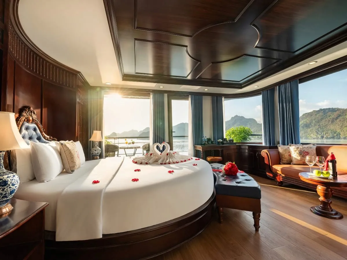 A luxury wood-paneled cruise cabin with a round bed decorated with towel swans and red petals, a sofa, a table with wine glasses and a bottle, and large windows showing sunlit limestone karst hills over the water.