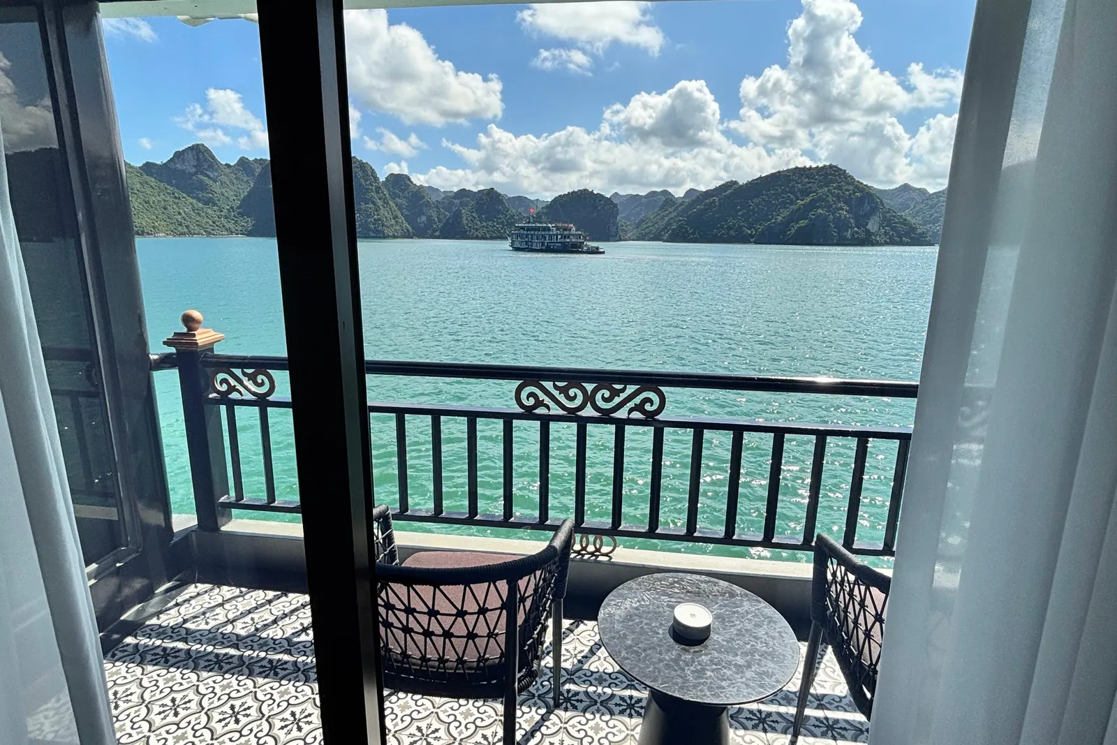 View through open sliding doors from a cabin onto a private balcony with two chairs, a small round table and patterned tile floor, overlooking turquoise water, limestone karst islands and another cruise boat under a partly cloudy sky.