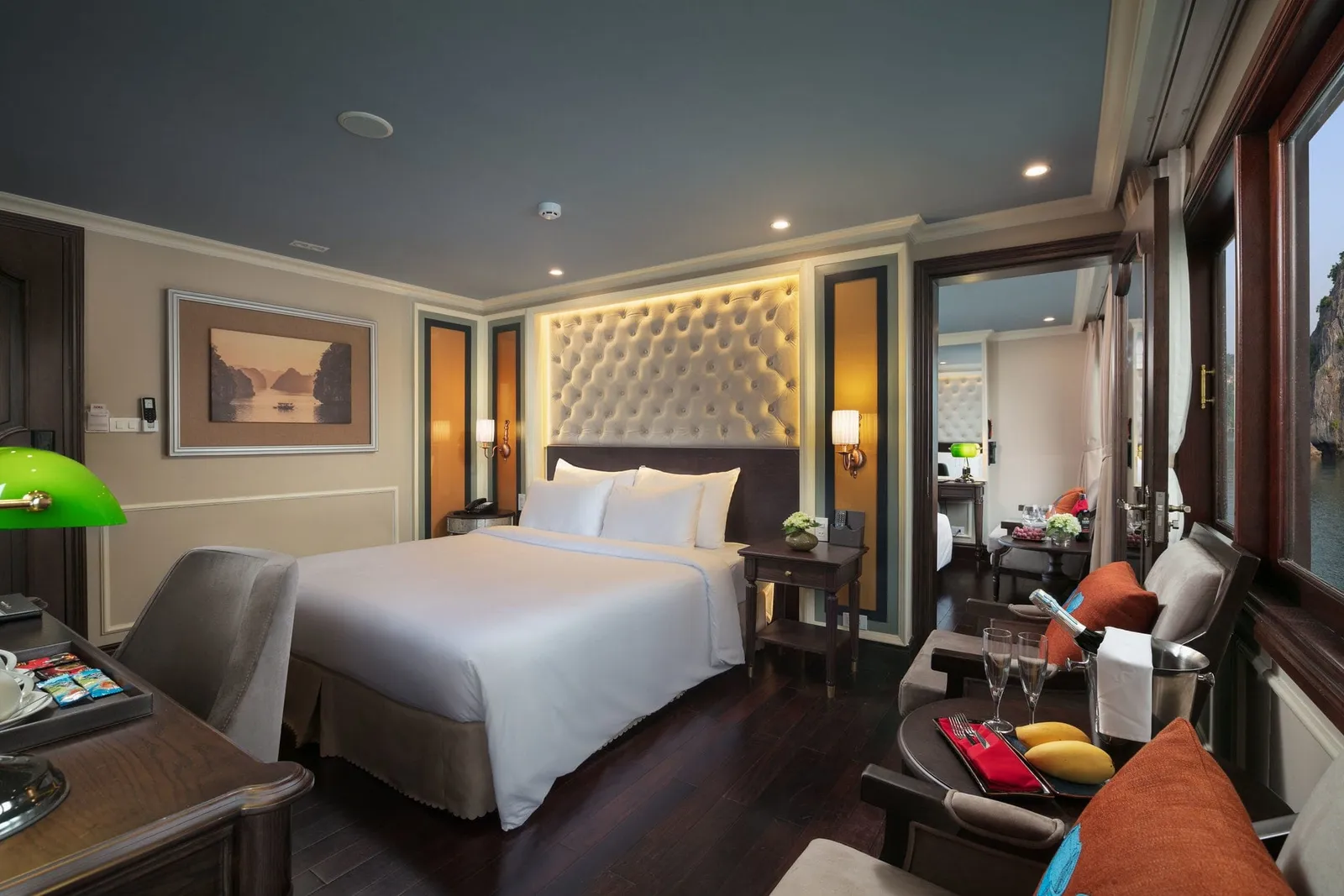 A cabin with a white double bed, tufted headboard, dark wood floor and furniture, a green desk lamp, and a champagne bucket with mangoes on a table by a window showing a limestone karst and water.
