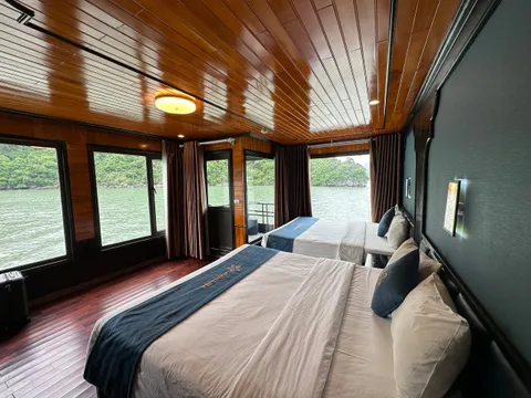 A wooden cabin interior with two beds, large windows showing water and karst islands outside, and text embroidered on the bed runners.